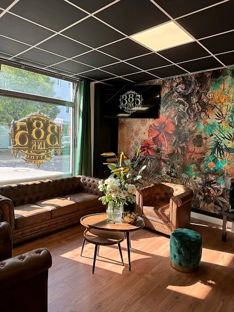 Lounge-Bereich mit Chesterfield-Sofas in Cognac, türkisem Velvet-Pouf, warmem Holzboden und goldenem 385-INK-Wappen-Logo am Fenster.