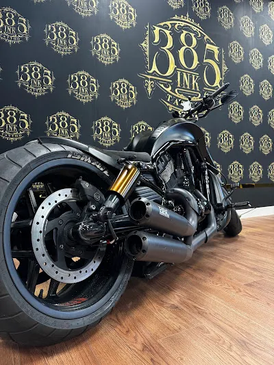 Schwarzes Custom-Motorrad mit goldenen Federbeinen vor einer Wand mit goldenem 385-INK-Wappen-Logo als Wiederholungs-Tapete.