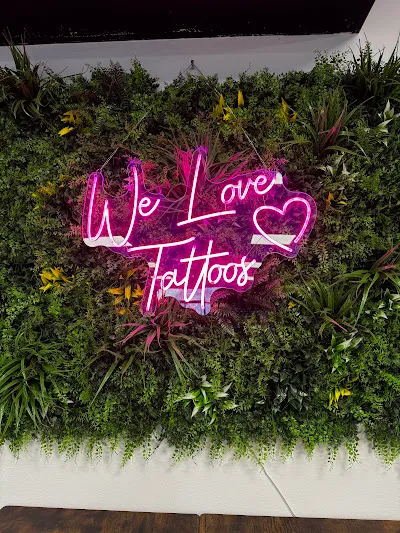 Pink-Neon-Schriftzug ‚We Love Tattoos‘ mit Herzchen, davor eine üppige vertikale Greenwall aus Farn, Hängepflanzen und gelben Blüten.